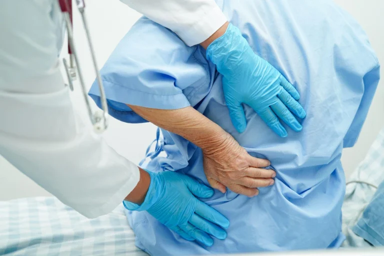 A doctor in blue gloves examining a patient's lower back, illustrating the initial consultation for joint and spine pain at Dr. Gadekar's clinic.